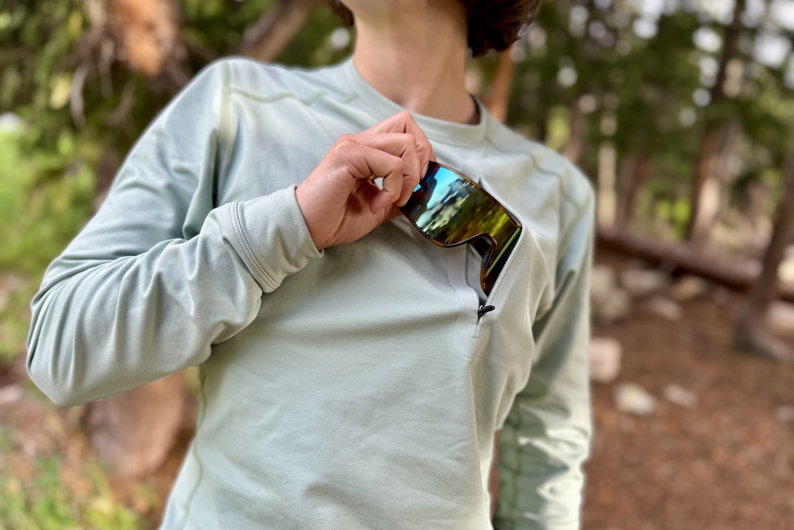 A hiker stands in the woods in a light blue base layer while pulling a pair of dope sunglasses out of her breast pocket