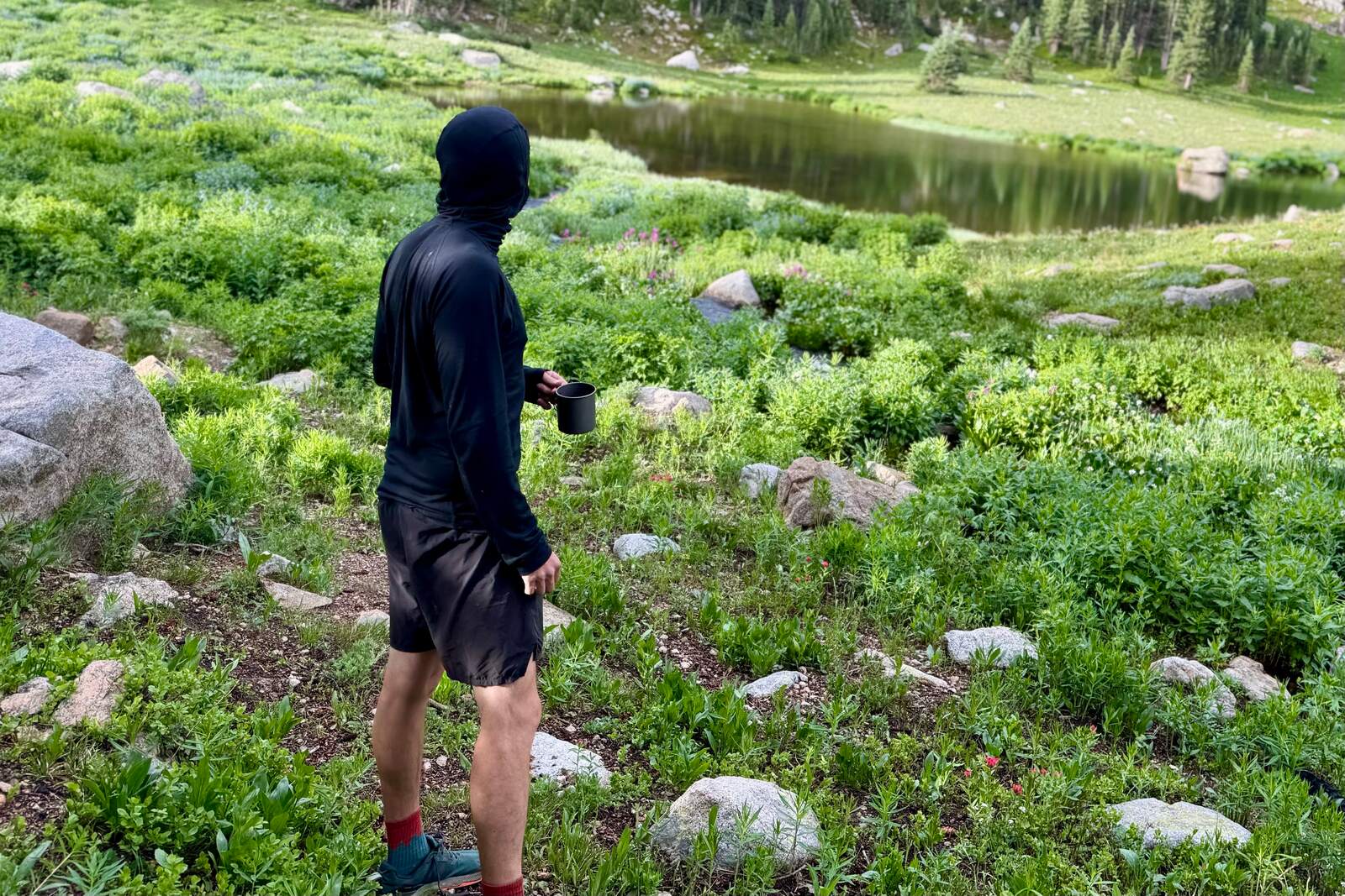a hiker in a black hooded base layer holds a mug while he stands in a field, gazing at an alpine lake