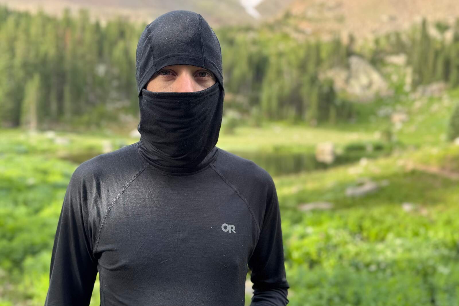 a hiker in a hooded and masked black base layer stands in front of the camera with a blurry alpine mountain background
