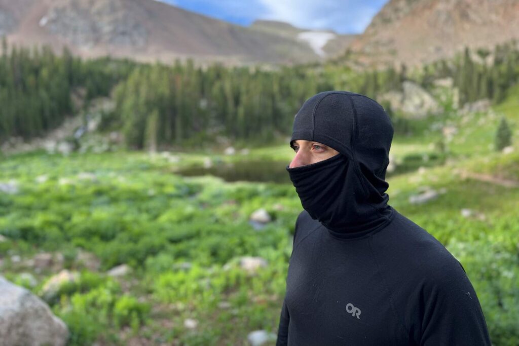 a hiker in a black bas layer with a hood and face mask stands in an alpine environment staring across the frame