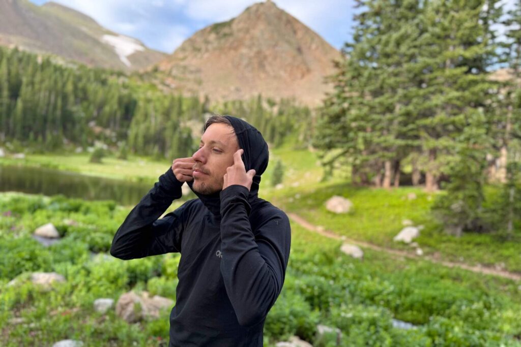 a hiker in a black hooded base layer stands in an alpine environment and raises the hood on his shirt