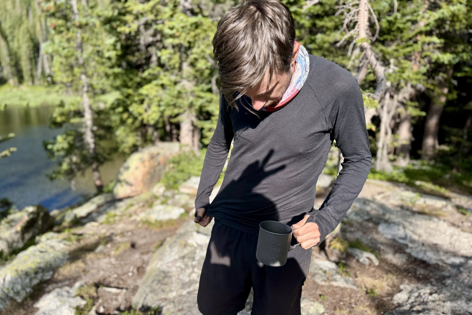 a hiker stands in the woods with a coffee mug, looking down at his gray base layer