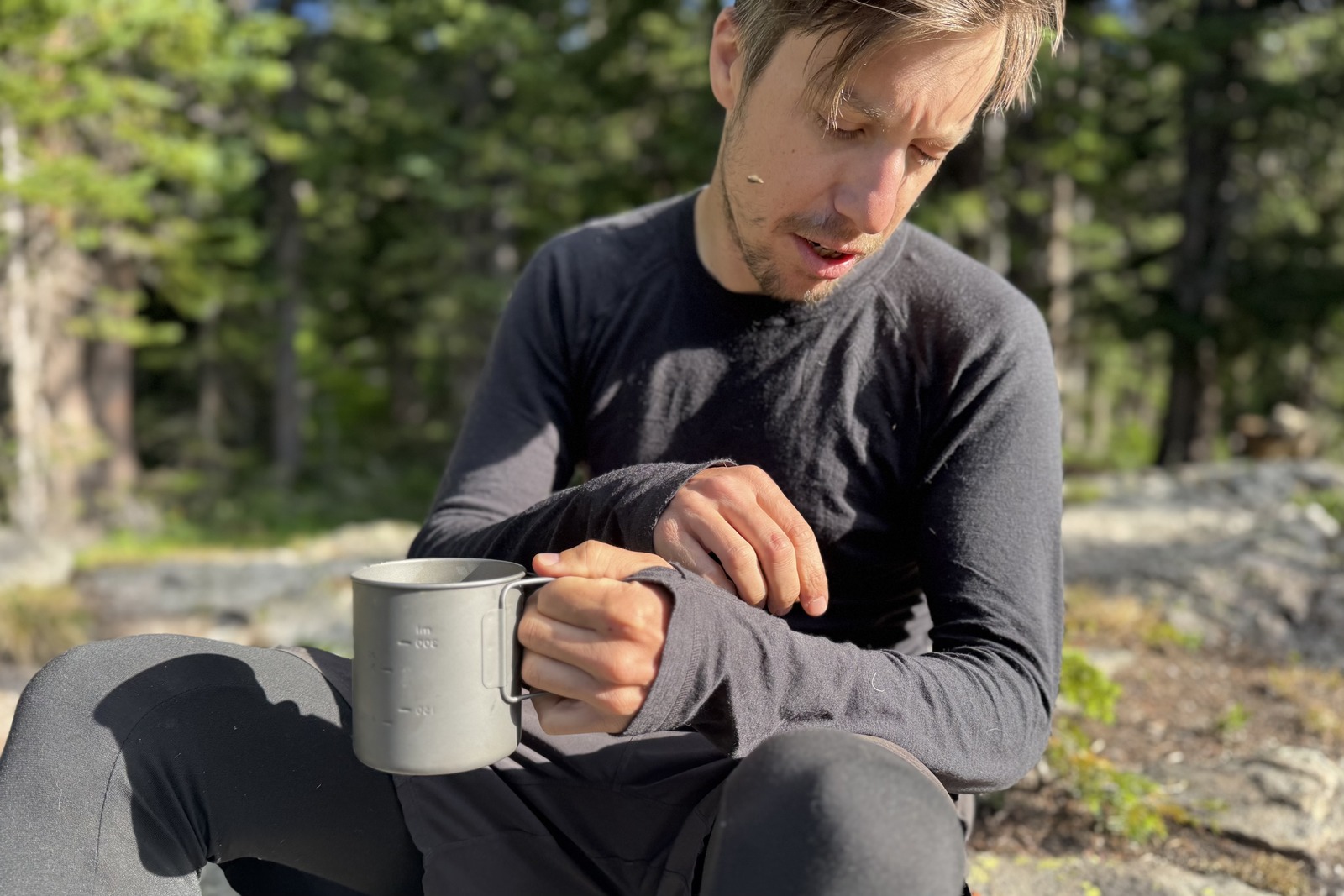 a hiker sits on a rock with a coffee mug, looking at his gray base layer