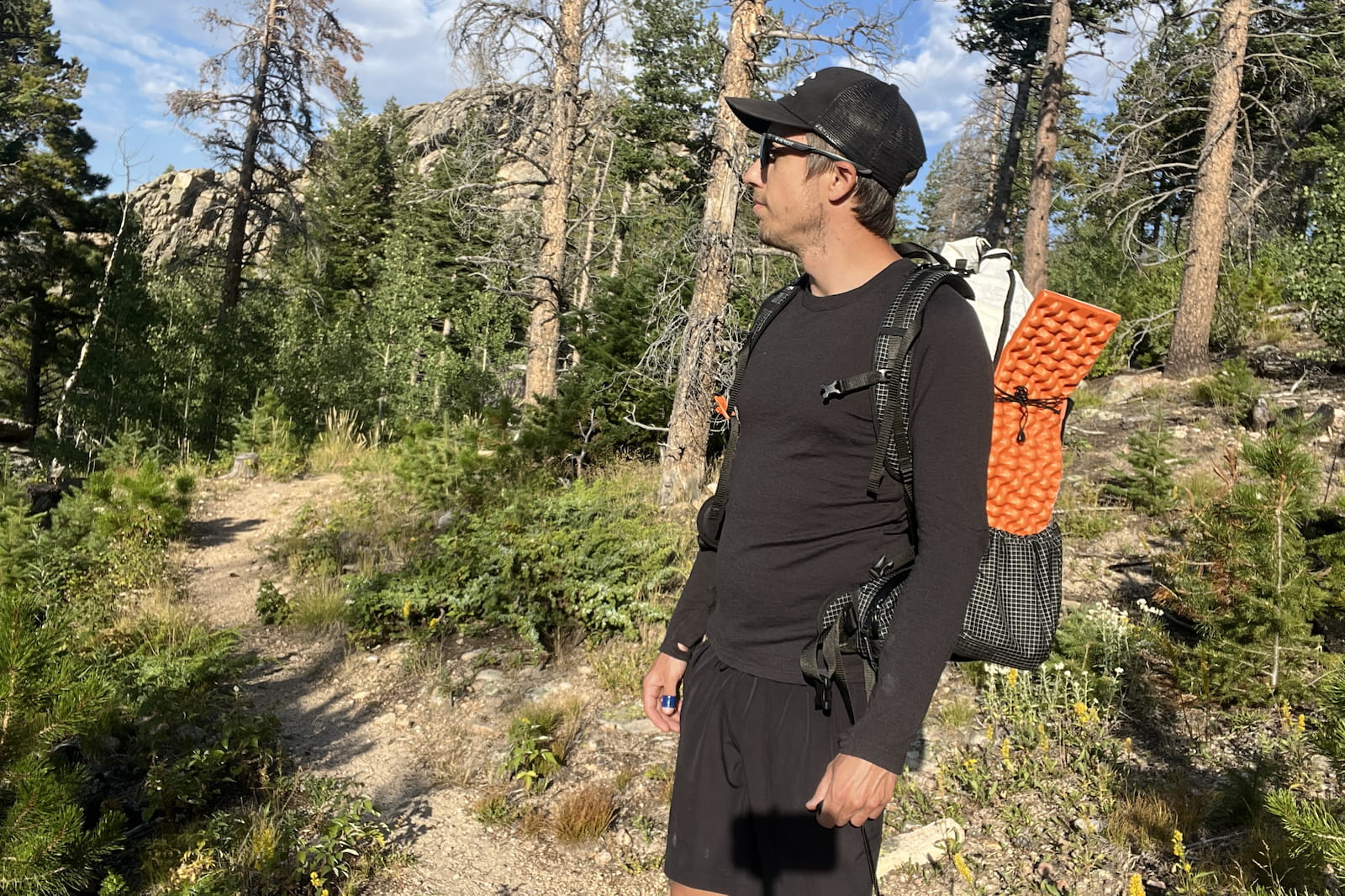 a hiker stands on a wooded trail in a black base layer, wearing a backpack