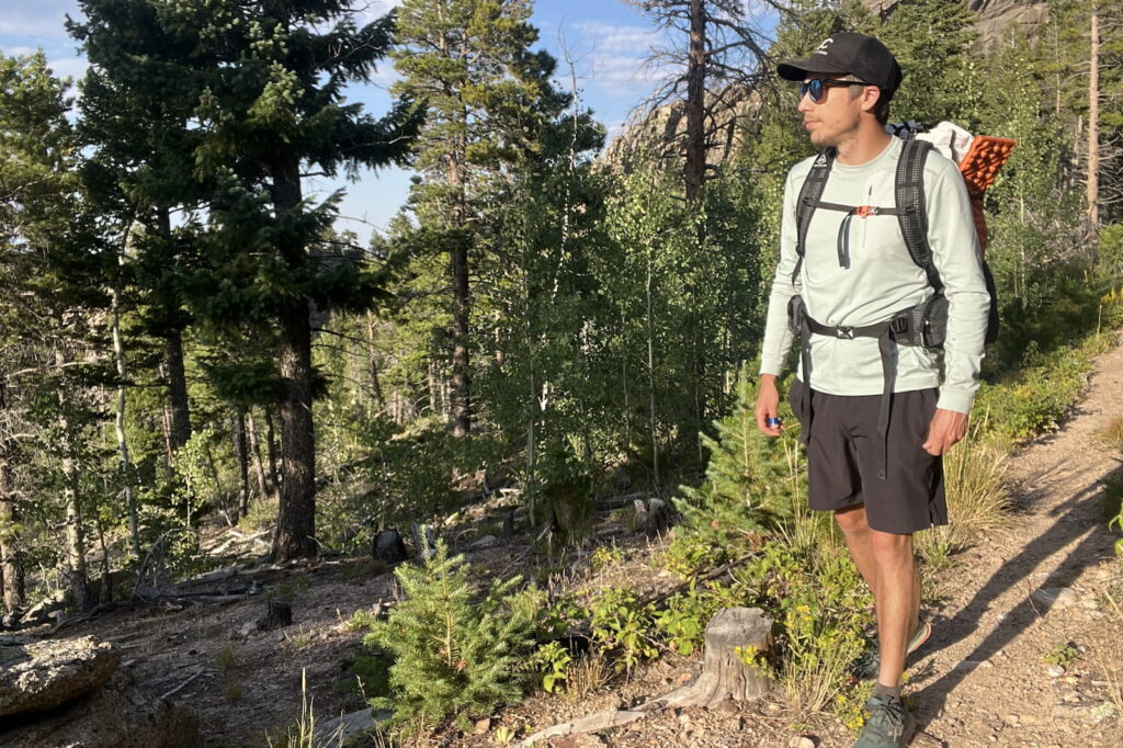 a hiker walks along a wooded trail wearing a light blue base layer and a backpack