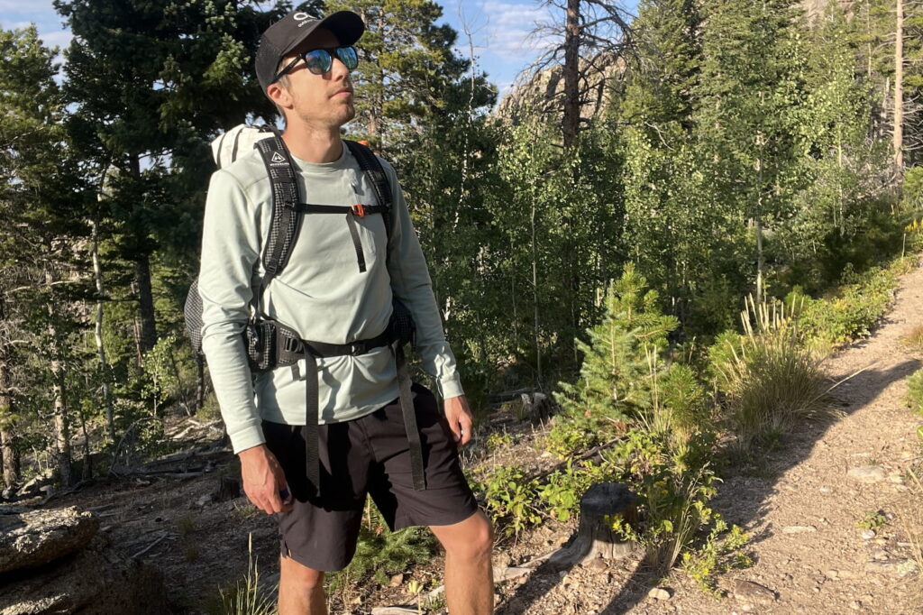 a hiker stands on a wooded trail wearing a light blue base layer and a backpack