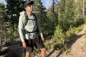 a hiker stands on a wooded trail wearing a light blue base layer and a backpack