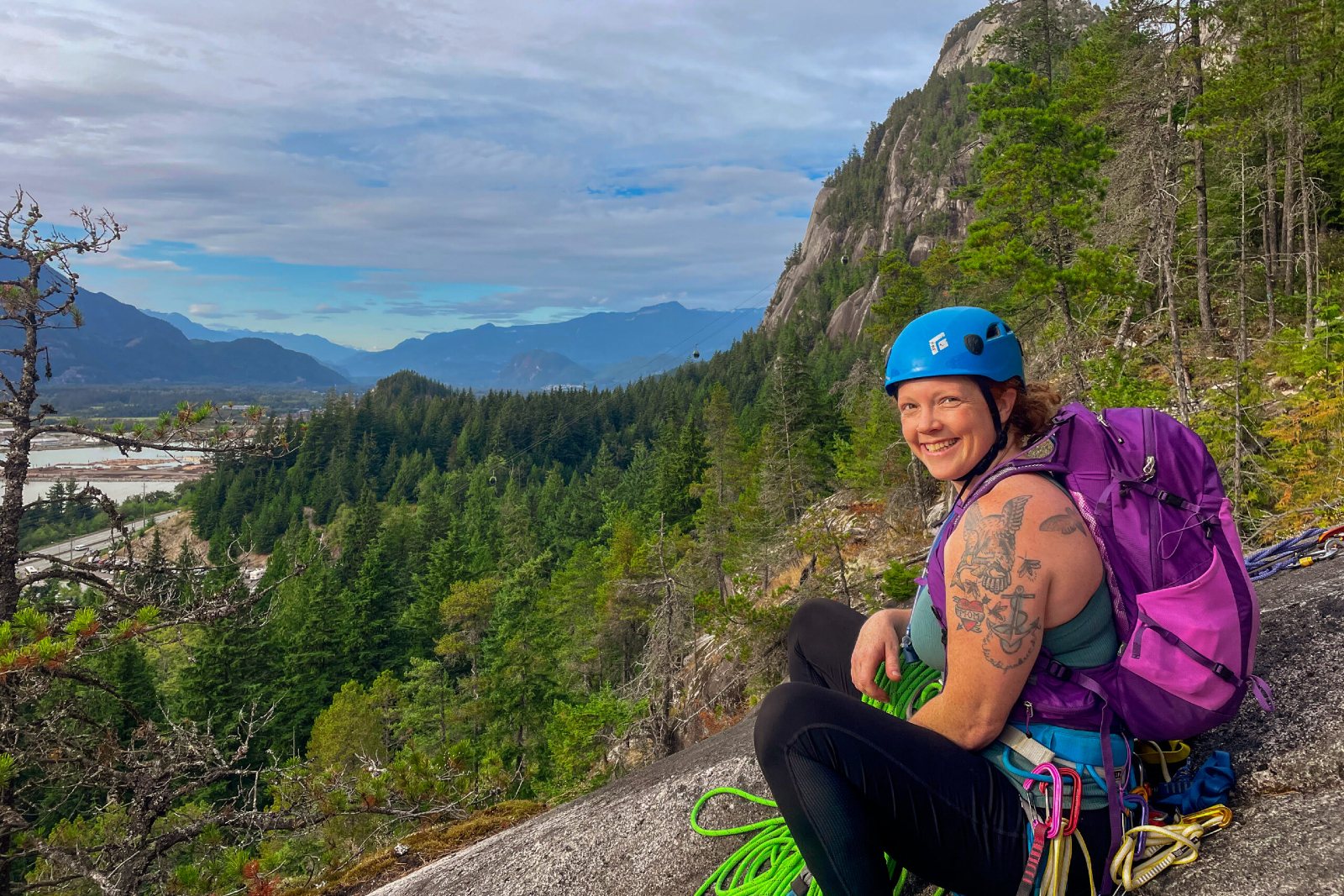 A person wearing a purple Deuter Speedlite Pro pack and a blue climbing helmet/harness smiles at the top of a rockwall. Mountain views of Squamish, BC are in the background.