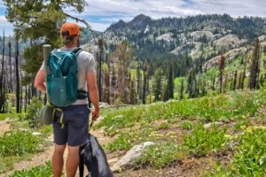 A person with a black dog looks out over a rocky landscape filled with pines. An EXPED brand pack is on their bag and a fly fishing rod is strapped to the side.