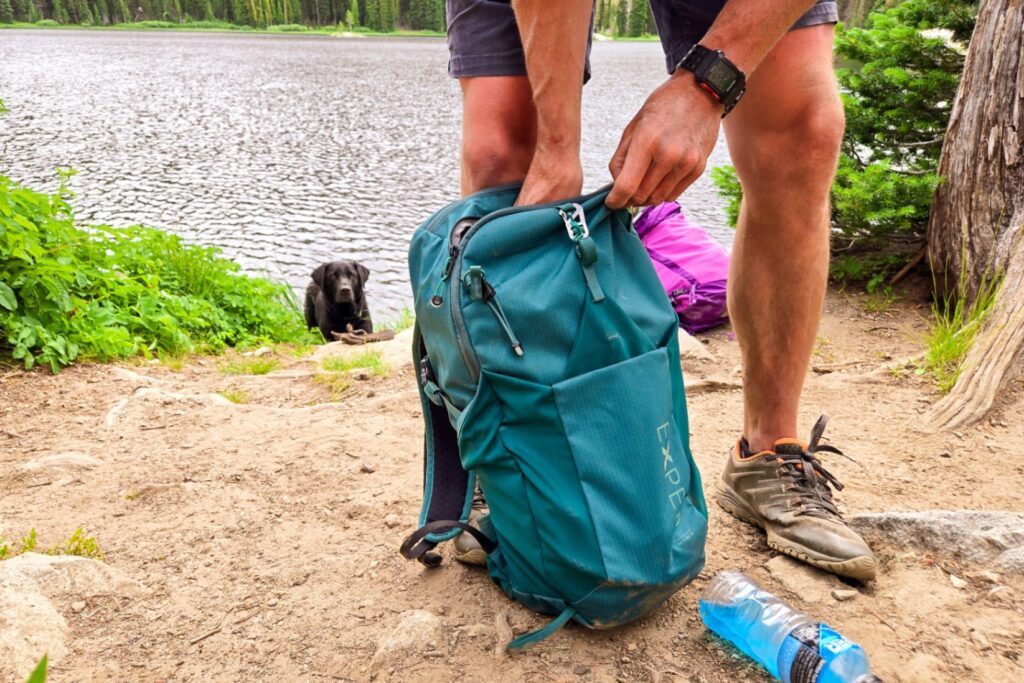 A person packs items into a green EXPED Skyline 20L Daypack. A black lab watches from the background.