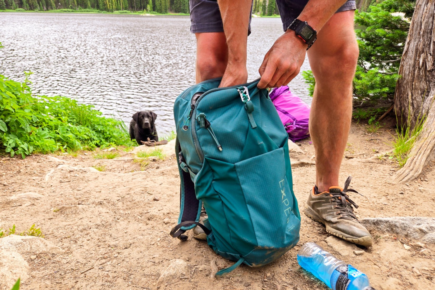 A person packs items into a green EXPED Skyline 20L Daypack. A black lab watches from the background.