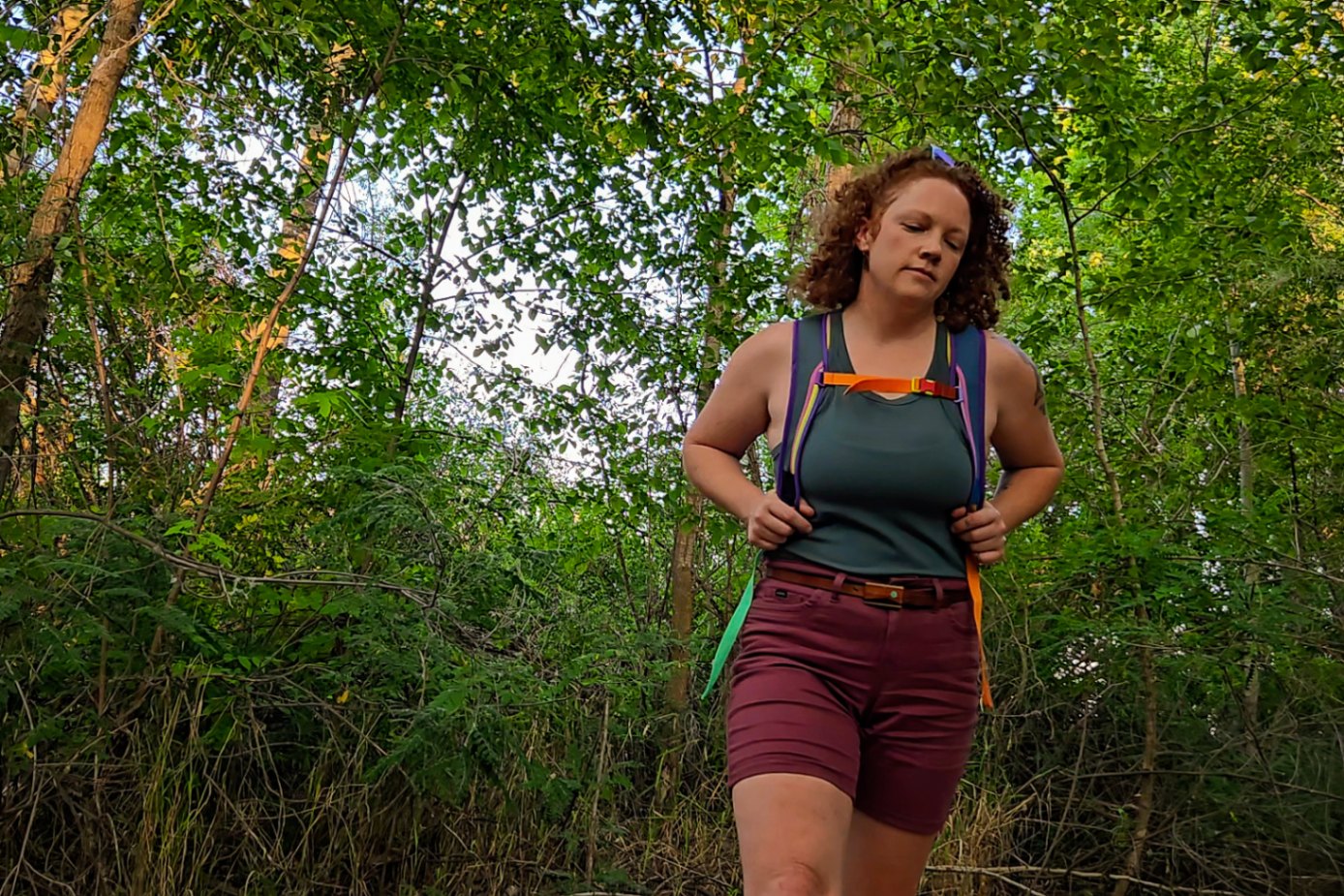 A person wearing a Cotopaxi Batac 16L daypack walks through a forested area. Wide mesh straps provide a comfortable fit.