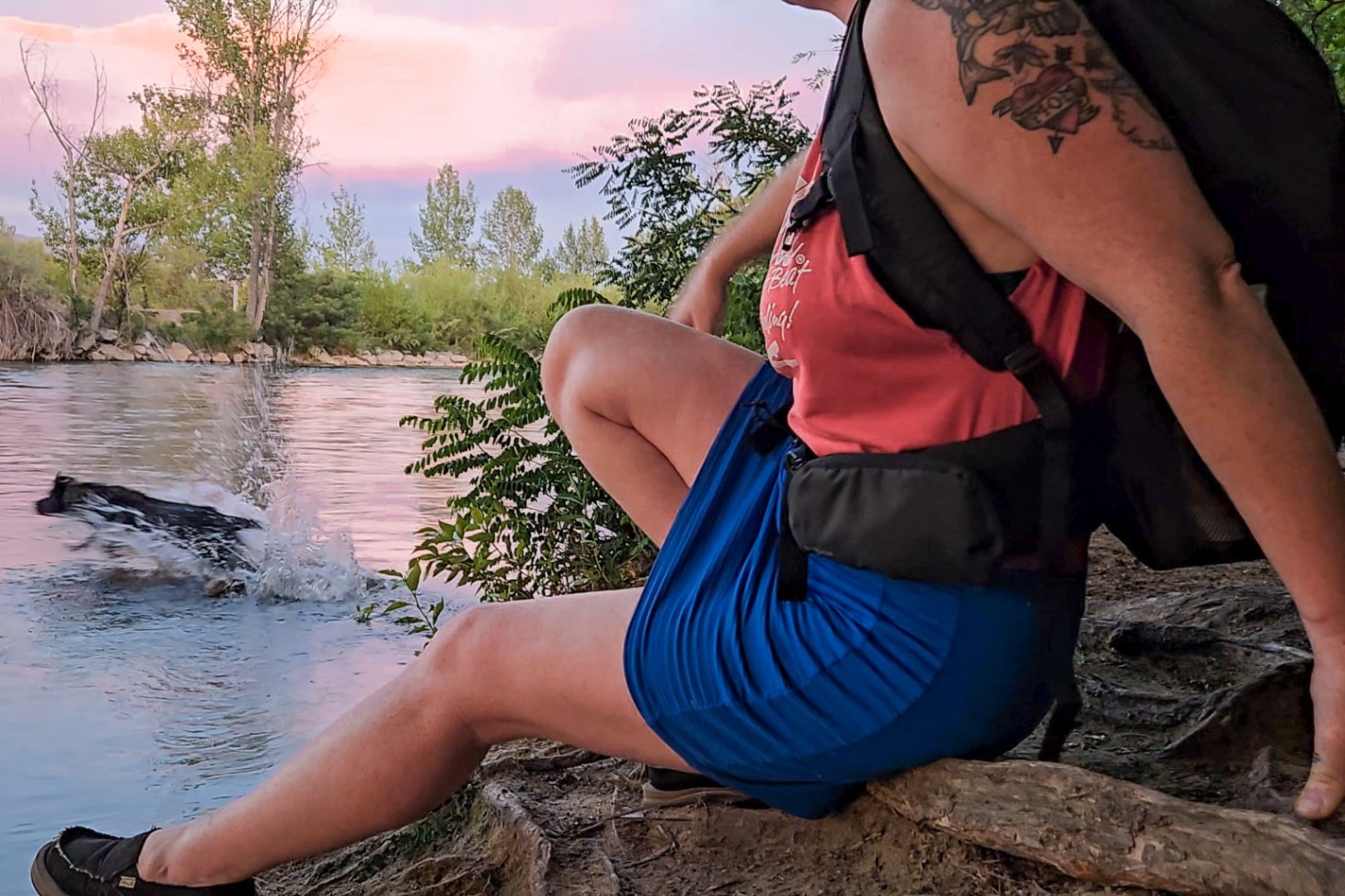 A person wearing a black daypack sits on a riverbank while a black dog jumps in the water. The backpack has adjustable shoulder straps and waist belt.