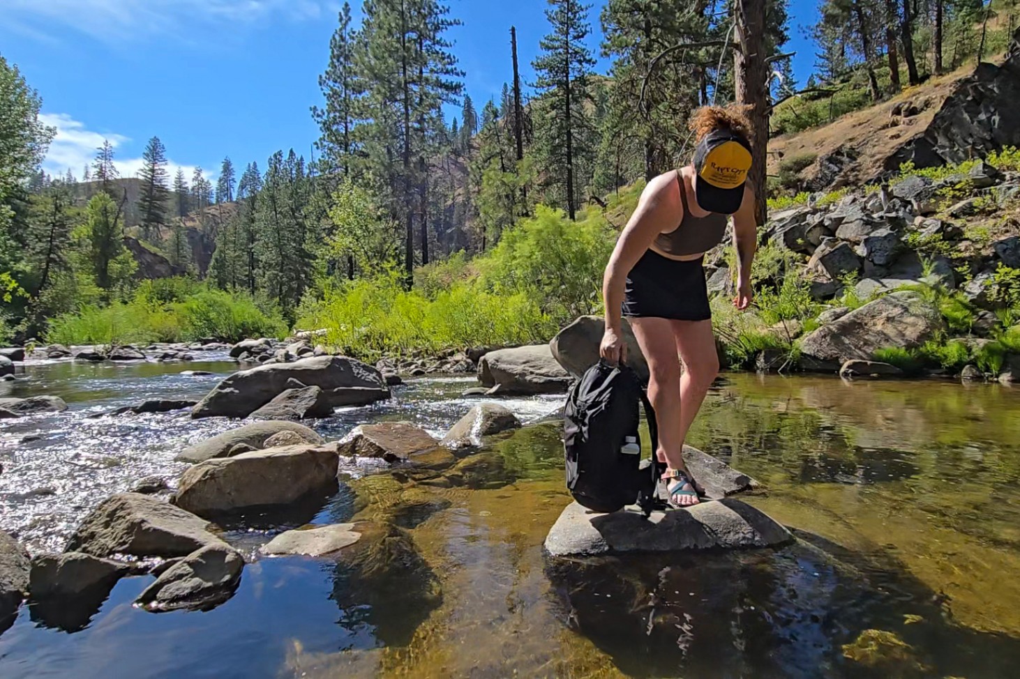 A person holding a black Decathlon Quechua daypack crosses a river. The side mesh pockets are displayed.