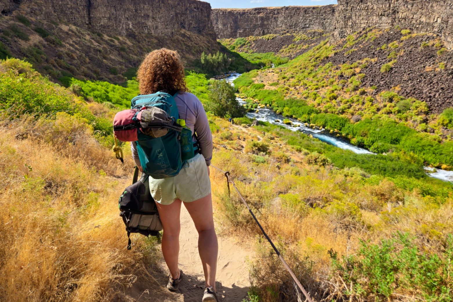 Person wearing a green EXPED Skyline 20L daypack hikes into a lush desert canyon carrying a PFD and a rescue bag strapped to the pack.