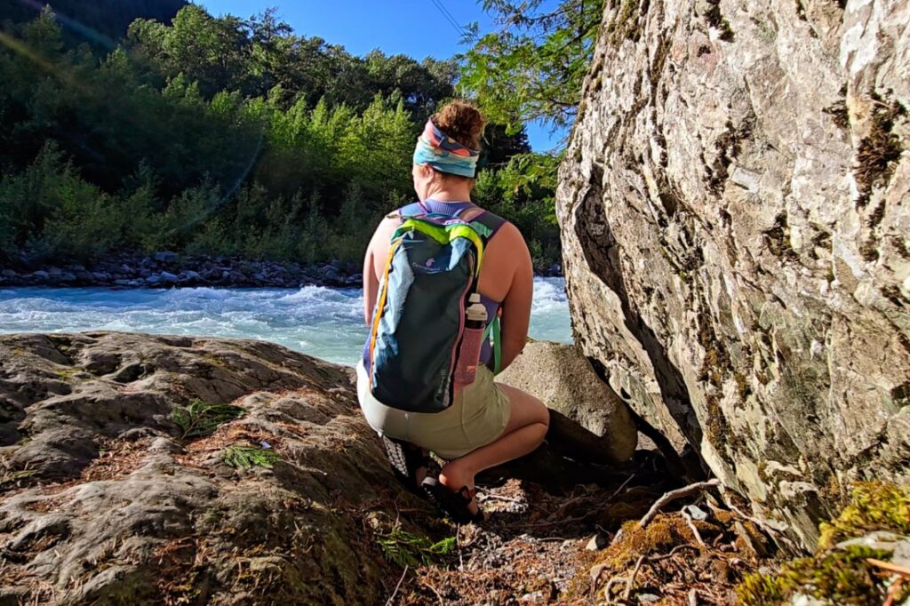 Person wearing a colorful Cotopaxi daypack squats by a riverside to watch the rapids.
