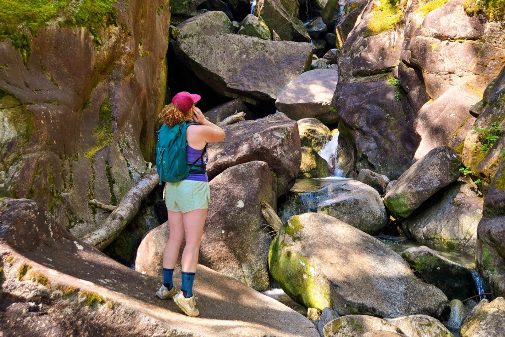 Person wearing a green EXPED daypack looks up at a rock jumble in a waterfall.