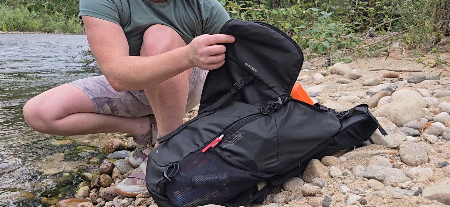 A person on a cobblestone riverbank opens a Decathlon Quechua MH100 20L day pack. The side view shows several items stashed in the mesh side pocket.