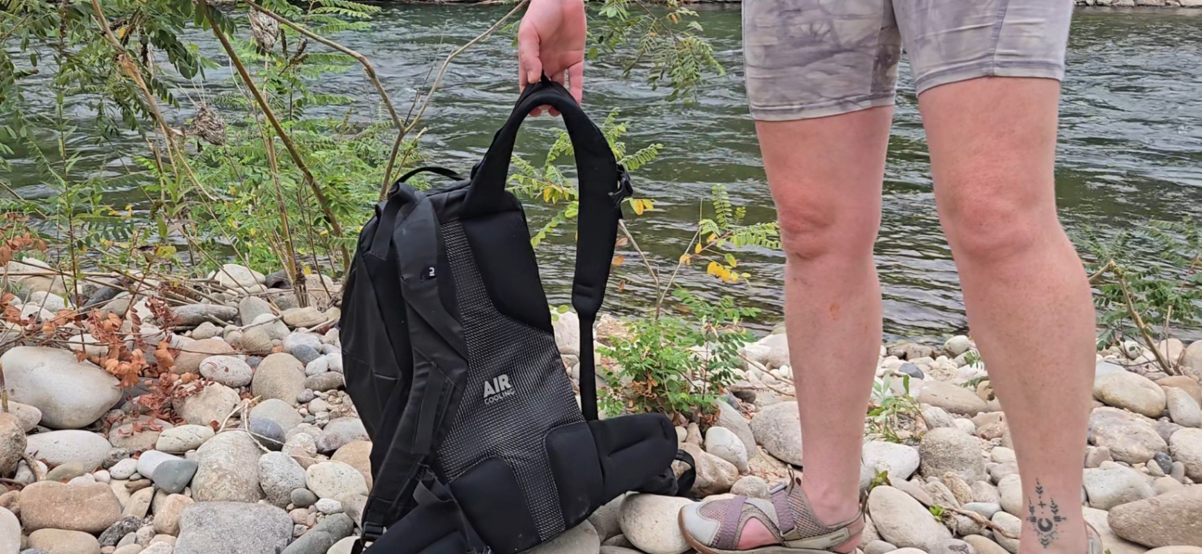 A person stands on a cobble riverbank and holds the Decathlln Quechua MH100 20L pack before they lift it to put it on. You can see the ample back padding and "Air Cooling" ventilation.