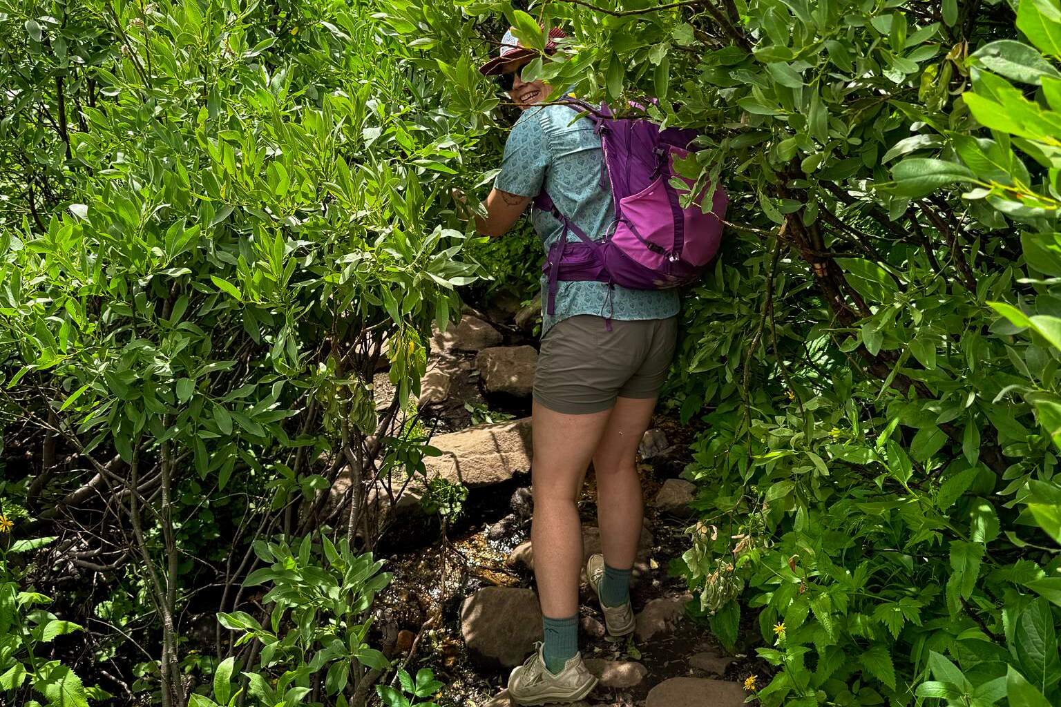 person wearing a purple Deuter Speedlite Pro pack grins as they push through bushes on an overgrown trail.