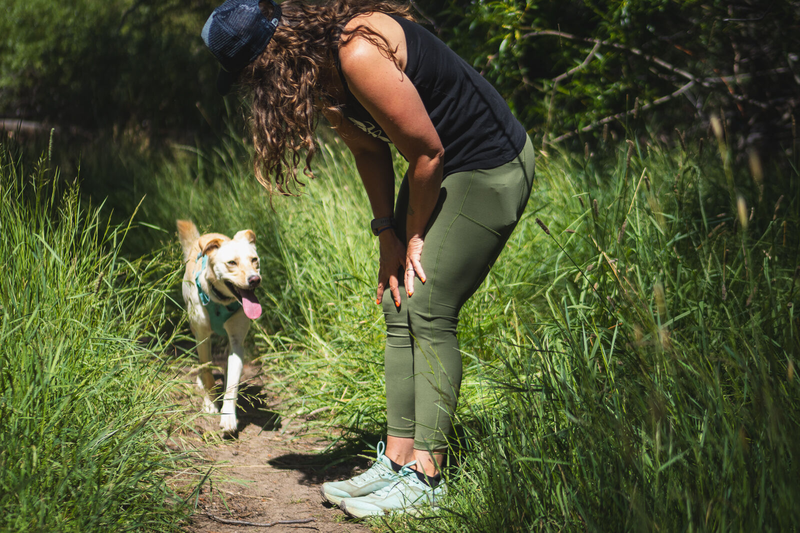 Women in green hiking leggings, curly hair, black tank top, teal dirty trail runner shoes, and black hat, bending at the waist with her hands on her thighs, looking at her dog who is running towards her on a dirt trail, surrounded by tall green grass. Dog has blonde fur and it's tongue is sticking out.