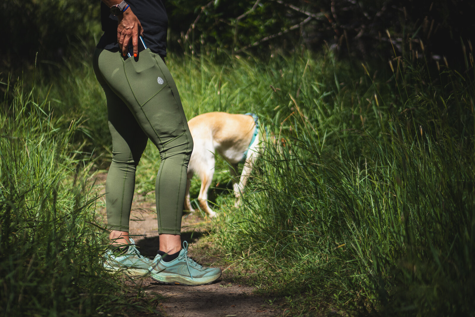 Women in green hiking leggings, pulling her phone out of the drop-in pocket, wearing dirty teal trail runner shoes, with a dog in the background in tall green grass