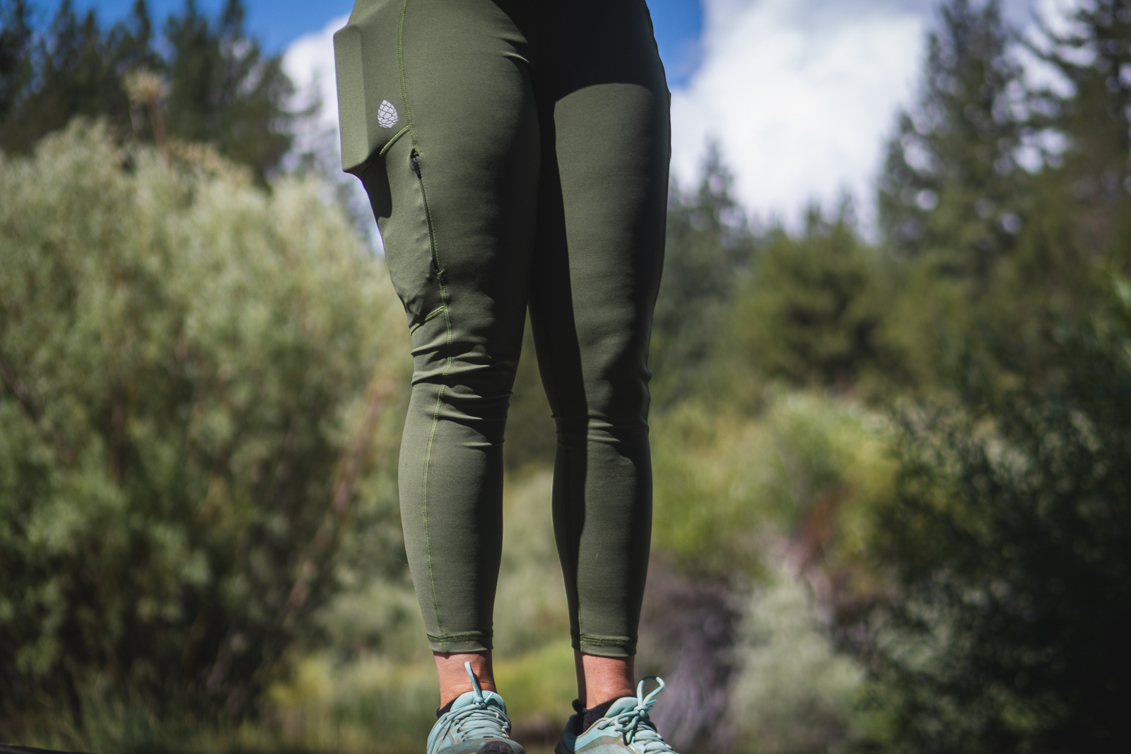 Women in green hiking leggings, camera zoomed in on legs showing a phone and other items in pockets. Background is green plants and trees, and a little blue sky with clouds.