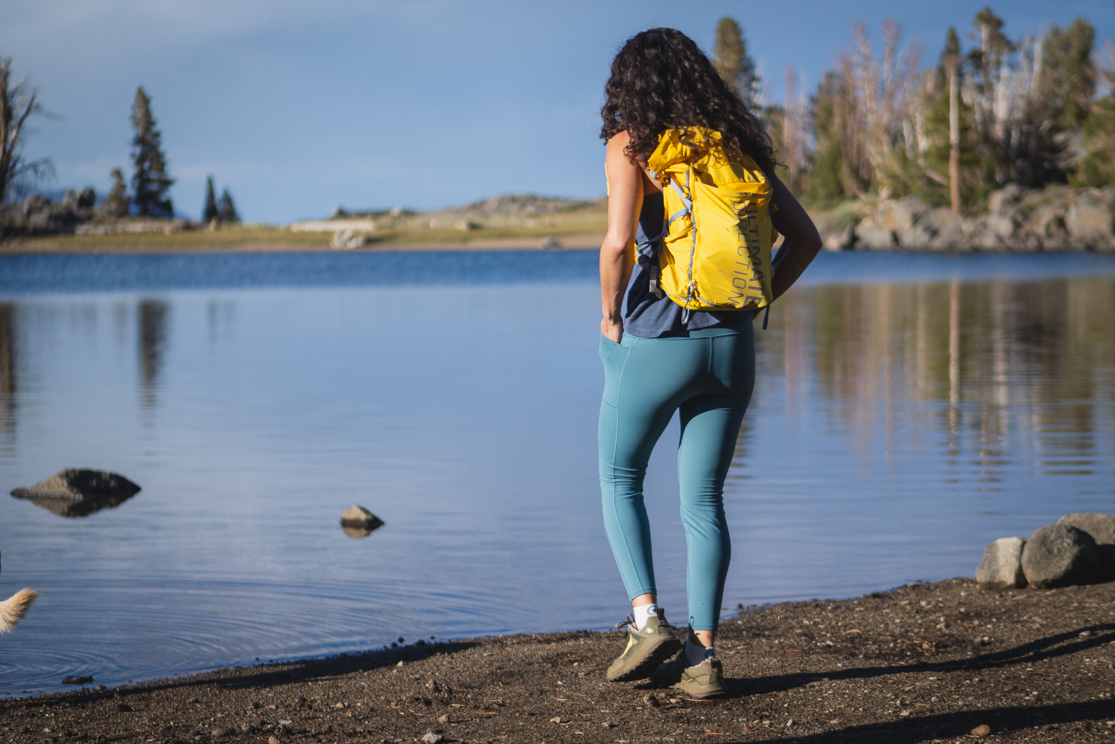 Woman with curly hair and sunglasses hiking in blue leggings, a dark blue tank top, and a yellow day pack. She's walking next to a reflective lake with the opposite shore showing dirt and pine trees.