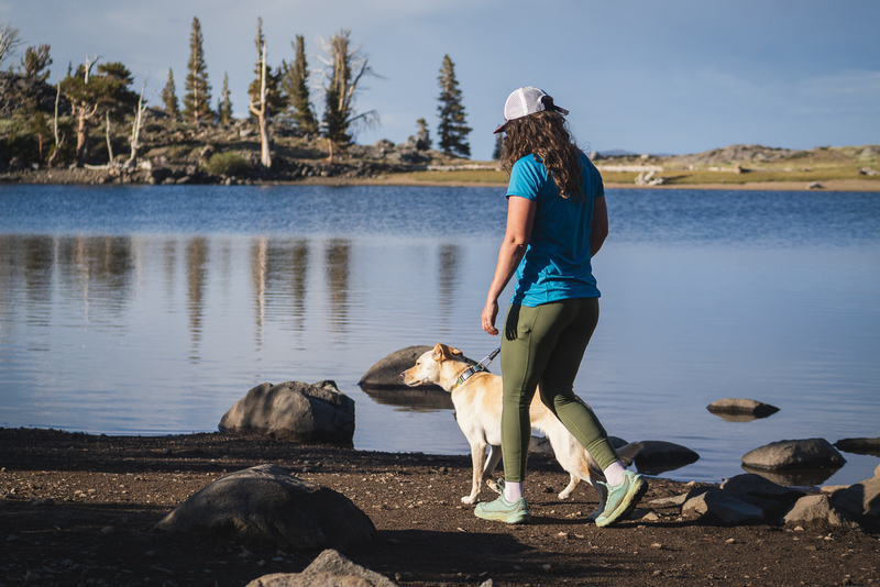 Women in green hiking leggings walking along side a lake with her dog. She's wearing a blue short and the dog has blonde fur. There are pine trees on the opposite side of the lake and some rocks in the foreground.