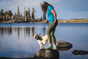 Women in green hiking leggings, standing on a rock in a lake, with her dog, with trees in the background on the other side of the lake.