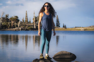 Woman with curly hair and sunglasses hiking in blue leggings, a dark blue tank top. She's standing on a rock with a reflective lake behind her, and pine trees in the distance.