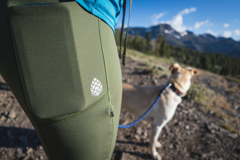 Women in green hiking leggings, showing the Stio logo, with a phone in the pocket, and a dog in the background, plus a trail, trees and a mountain