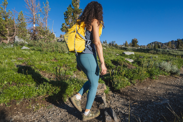 Woman with curly hair and sunglasses hiking in blue leggings, with a yellow day pack, a dark blue tank top. Background is green, with pine trees and granite mountains. Lighting is just before sunset. She's walking down a dirt trail.
