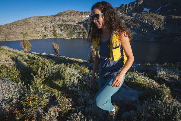 Woman with curly hair and sunglasses hiking in blue leggings, with a yellow day pack, a dark blue tank top. She's climbing uphill, with a dark blue lake behind her that is framed by tall granite mountains. There are green plants by her feet.