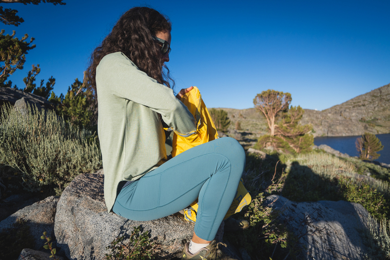 Woman with curly hair and sunglasses hiking in blue leggings. She's pulling something out of her yellow backpack. She's wearing a greyish green long sleep. Sitting on a granite rock, with a tree in the background.
