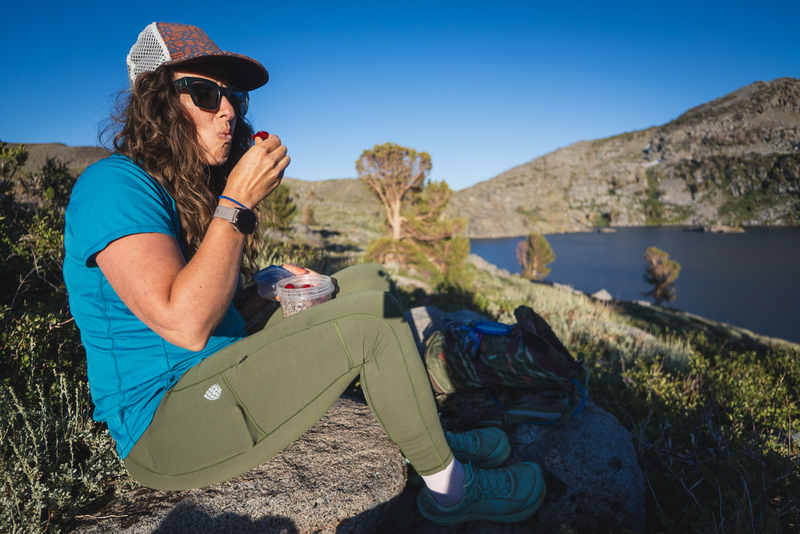 Women in green hiking leggings with curly hair, sunglasses, and a white and brown hat on, sitting on a rock, having a snack, with a lake, a tree and some hills in the background.