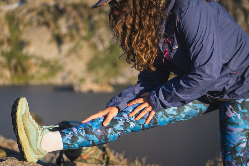 Woman wearing bright blue leggings in a camouflage like pattern on a hike, with one foot up on a rock, stretching her calf and hamstring. She has curly hair and is wearing a purple rain jacket.