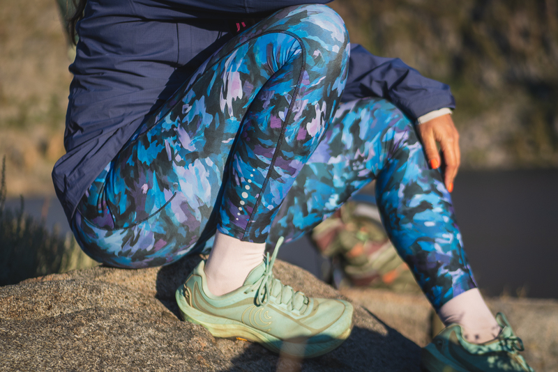 Woman wearing bright blue leggings in a camouflage like pattern on a hike. She's sitting on a rock with knees bent and tucked in tight to her body. She's wearing light teal trail runners.