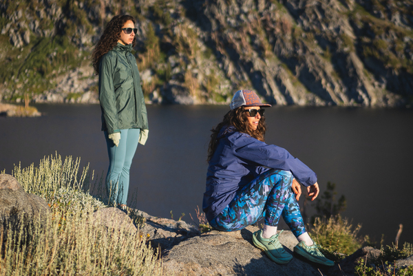 Woman wearing bright blue leggings in a camouflage like pattern with a purple rain jacket, sunglasses and a hat. She's sitting in the foreground, with knees bent and arms resting on her knees. Her friends is standing behind her, closer to the lake in the background. She's wearing lighter blue leggings, no pattern, with a green rain jacket, and sunglasses. Both have curly hair.