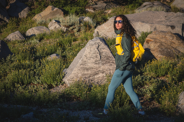 Woman with curly hair and sunglasses hiking in blue leggings, with a yellow day pack, a dark blue tank top and a green rain jacket. Background is green plants and granite boulders. She's looking out, seemingly toward a view that we cannot see.