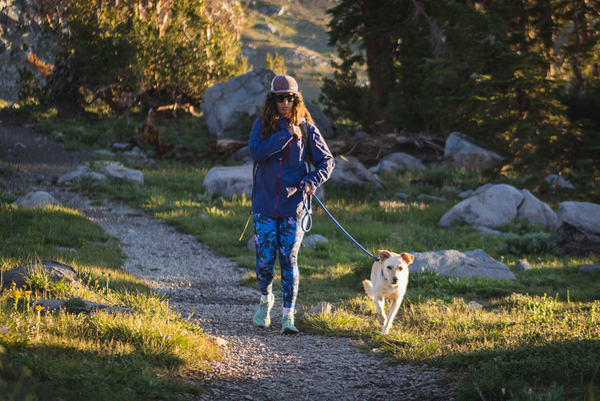 Woman wearing bright blue leggings in a camouflage like pattern on a hike. She's hiking towards the camera, with her dog leading the way attached by a leash. The dog has blonde fur. She's wearing a purple jacket and teal shoes, sunglasses and a hat. Along the trail are granite boulders, green grass and some pine trees.