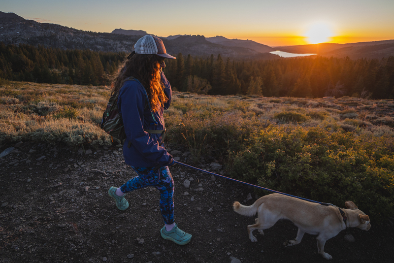 Woman wearing bright blue leggings in a camouflage like pattern on a hike. She's wearing teal train runners and a purple rain jacket and a hat. Her blonde dog is walking on the trail in front of her. In the background, the sun is setting behind some mountains, a lake, and some tall grass or flowers.