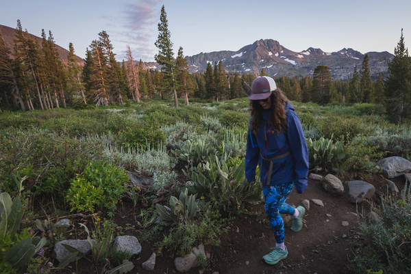 Woman wearing bright blue leggings in a camouflage like pattern on a hike. She has on a purple rain jacket, teal shoes, a hat and a daypack. In the background is a tall mountain with some snow on it, and a few pine trees.