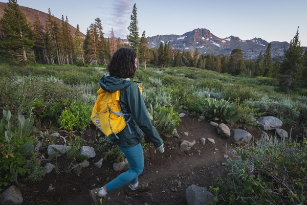 Woman with curly hair and sunglasses hiking in blue leggings, with a yellow day pack, a dark blue tank top and a green rain jacket. Background is green, with pine trees and granite mountains. Lighting is just before sunset. She's walking down a dirt trail.