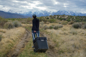 A person with their back to the camera pulls the YETI Roadie 48 cooler by its periscope handle down a path. There are mountains in the distance.