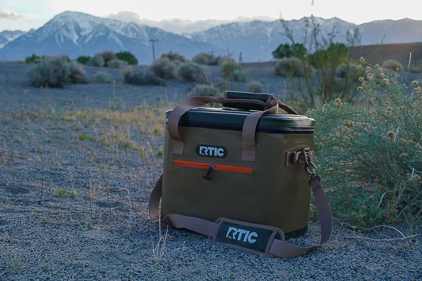 The RTIC Soft Pack cooler sits on sandy ground with mountains in the background.