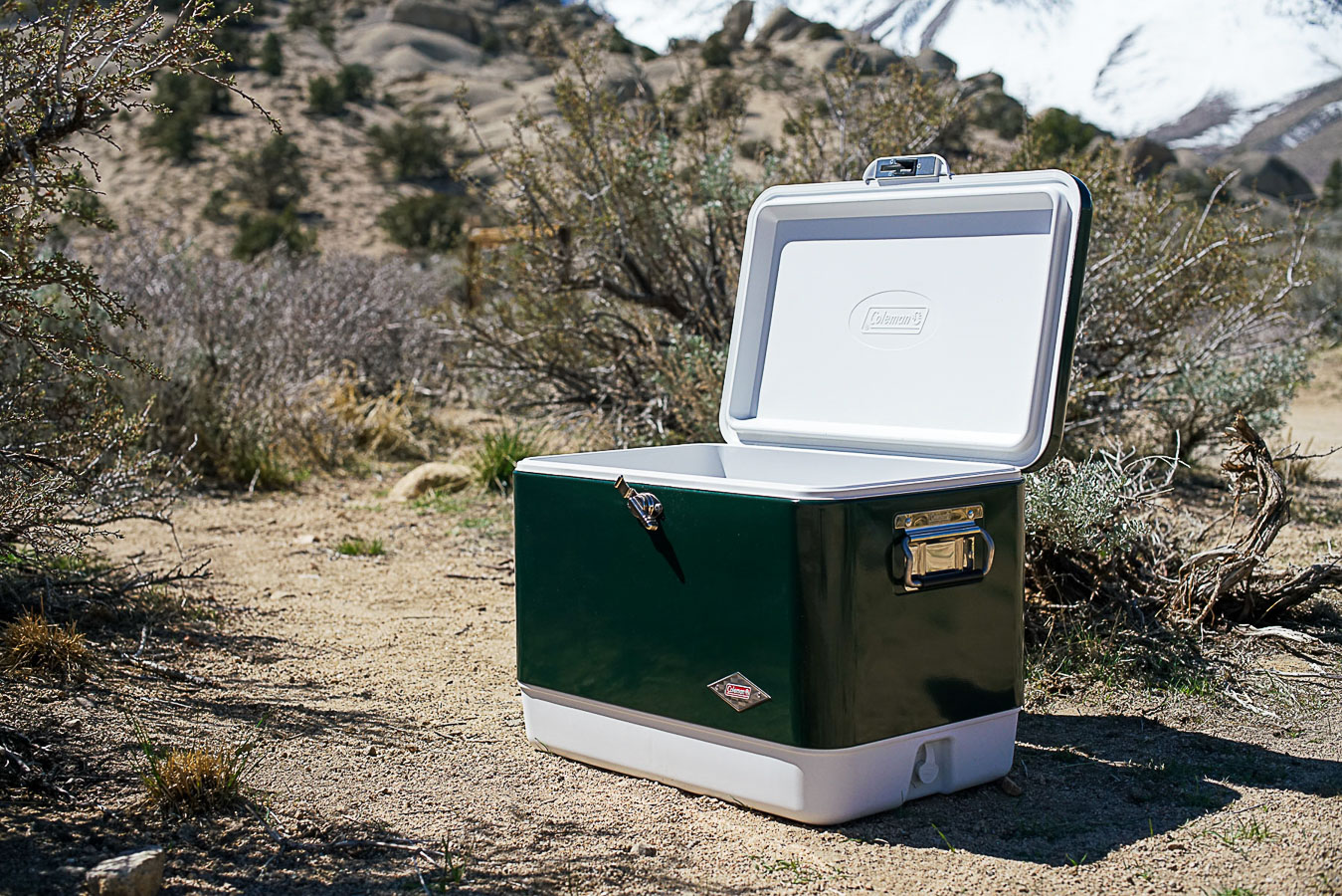 The Coleman Steel Belted cooler sits on the ground with sage brush and boulders in the background.