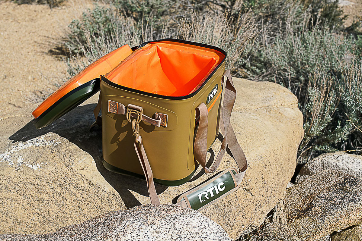 The RTIC Soft Pack cooler sits on a rock. The lid of the cooler is open, revealing an empty interior.