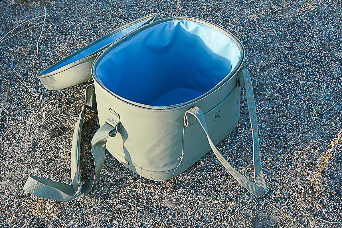 An open Hydro Flask Carry Out 20L cooler sits on sandy ground. The cooler is empty.