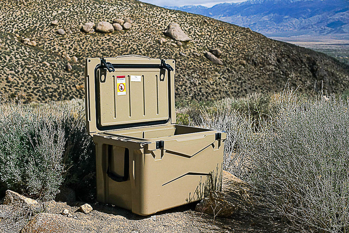 The XSpec 60QT cooler sits amongst sage brush with boulders in the background. The lid of the cooler is open.